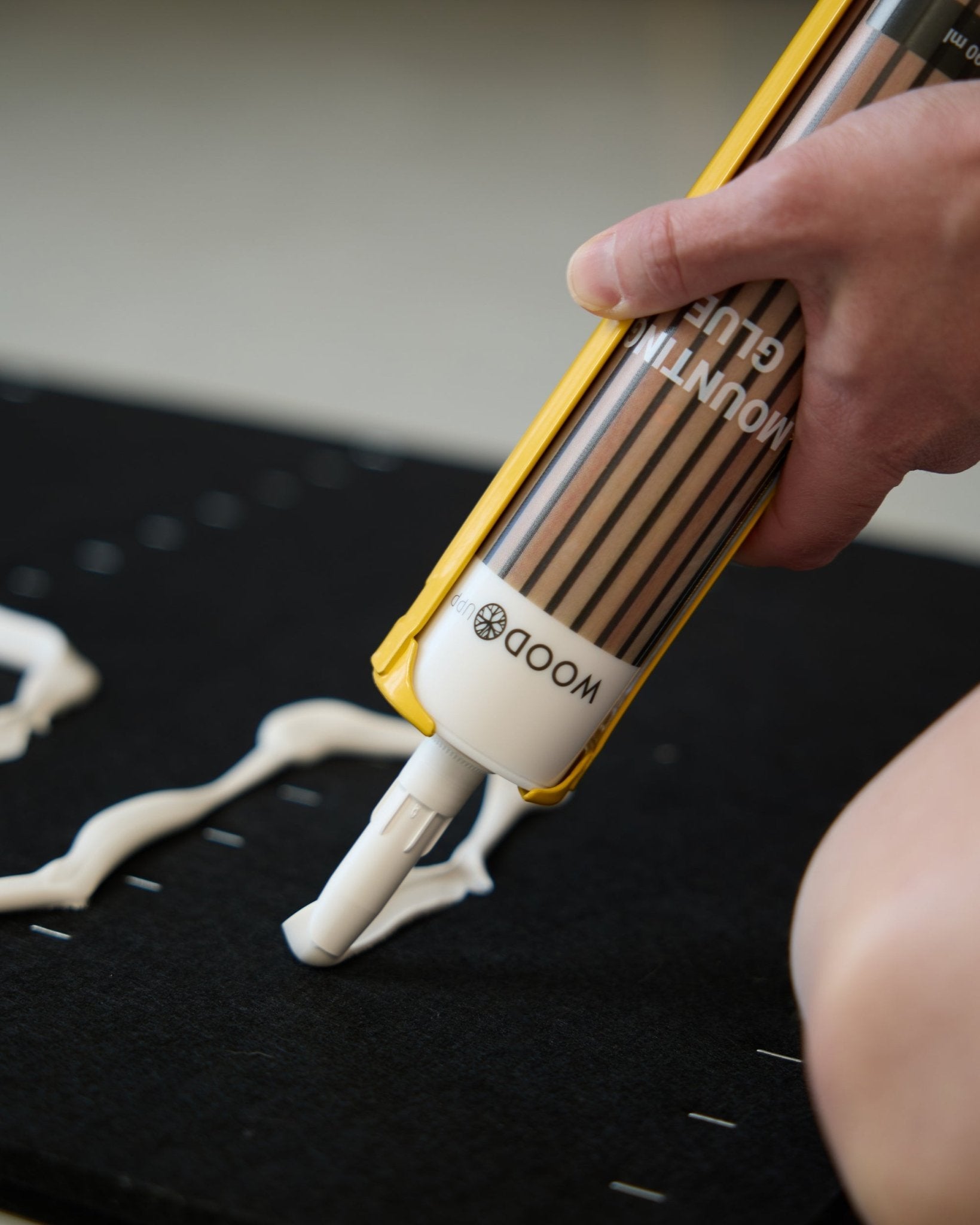 A hand applies wavy lines of WoodUpp Mounting Glue to a black surface in preparation for installing Akupanel acoustic wood panels.